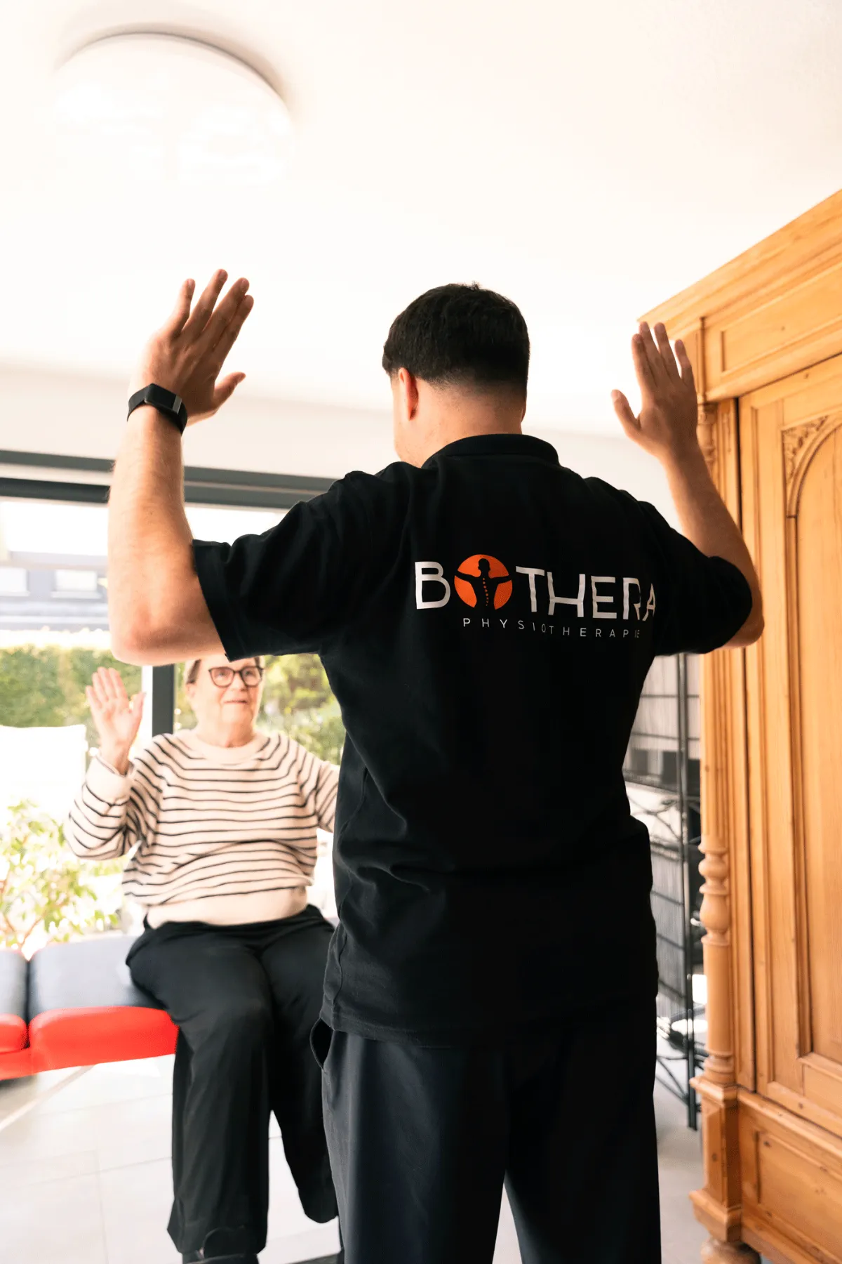 Ein Physiotherapeut zeigt einer älteren Patientin eine Übung mit erhobenen Armen.