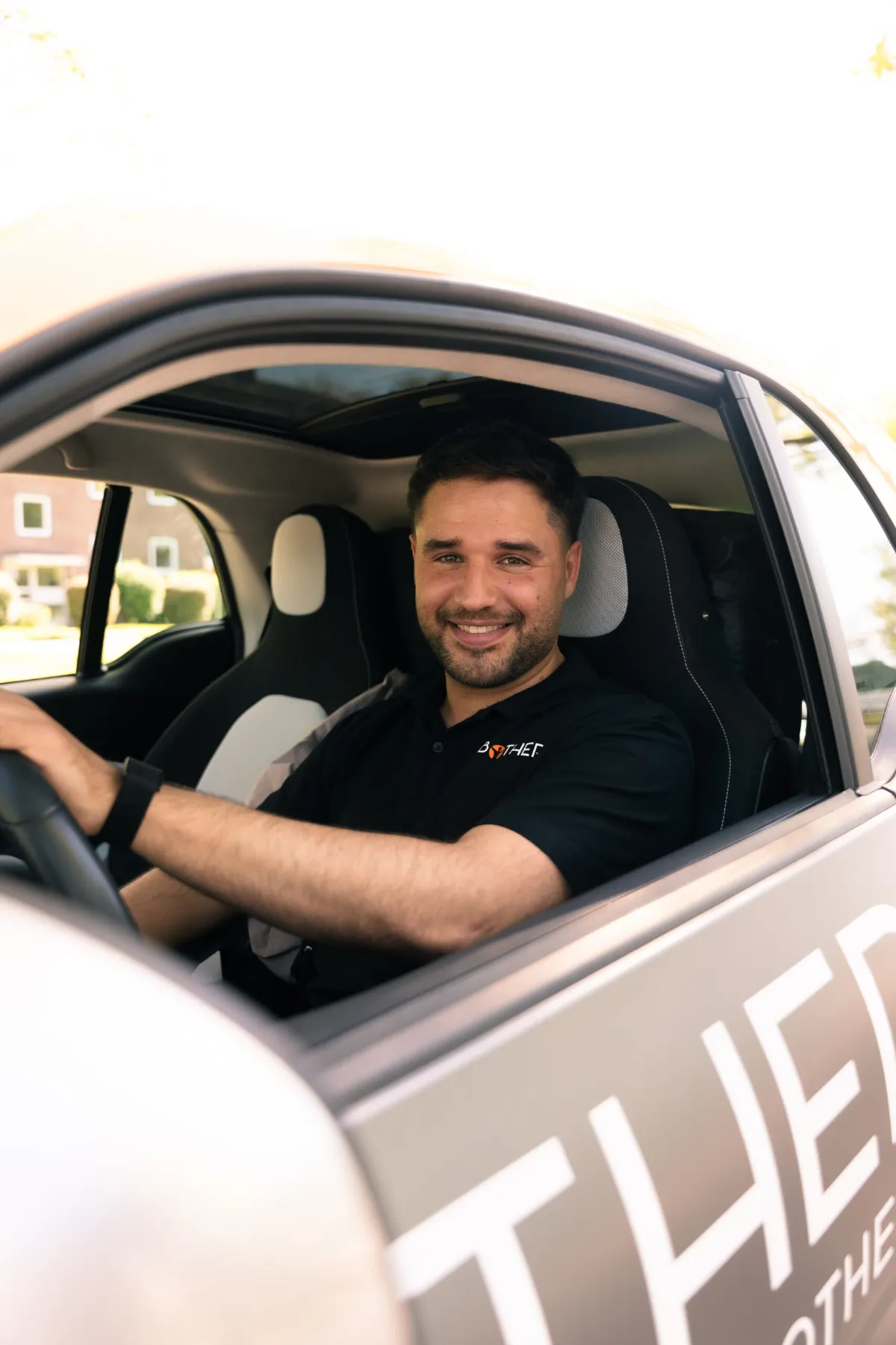 Ein lächelnder Bothera-Mitarbeiter im schwarzen Poloshirt sitzt am Steuer eines Firmenwagens.